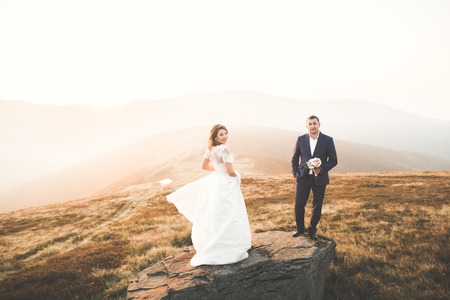 Young newly wed couple, bride and groom kissing, hugging on perfect view of mountains, blue skyの写真素材