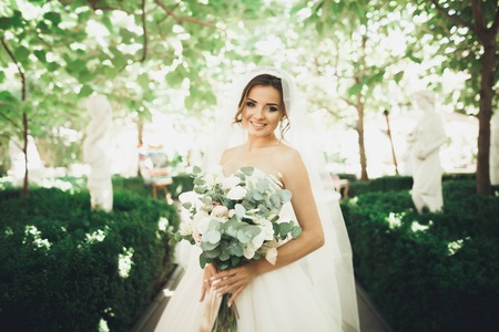 Beautiful brunette bride in elegant white dress holding bouquet posing neat treesの写真素材