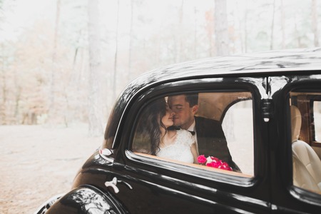 Stylish wedding couple, bride, groom kissing and hugging near retro car in autumnの写真素材
