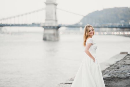 Beautiful brunette bride in elegant white dress holding bouquet posing near riverの写真素材