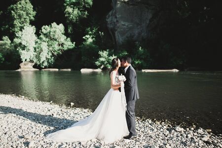 Elegant stylish happy blonde bride and gorgeous groom on the background of a beautiful river in the mountains.の写真素材