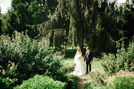 Stylish couple of happy newlyweds walking in the park on their wedding day with bouquetの写真素材