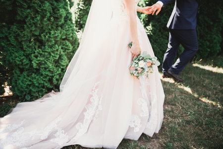 Stylish couple of happy newlyweds walking in the park on their wedding day with bouquetの写真素材