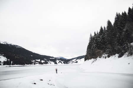 Romantic young couple looking away, back view, lake and winter mountains on backgroundの写真素材