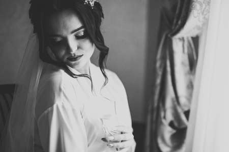Beautiful and lovely bride in night gown and veil sitting and drinking champagneの写真素材