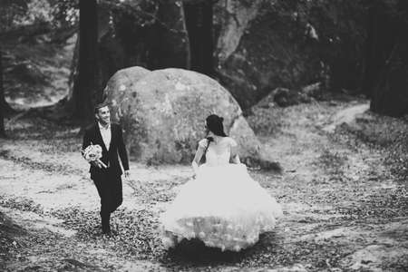 Stylish couple of happy newlyweds. Bride running from groom in the park on their wedding day with bouquetの写真素材