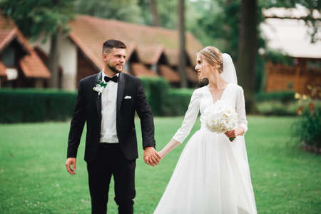 Stylish couple of happy newlyweds walking in the park on their wedding day with bouquetの写真素材
