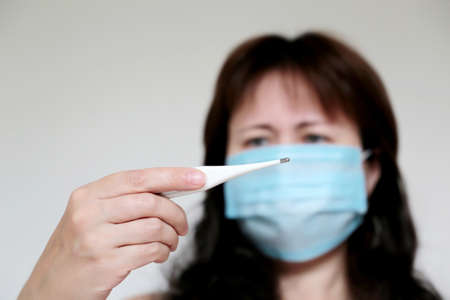 Coronavirus symptoms, woman in medical face mask measures body temperature. Worried girl looks at...