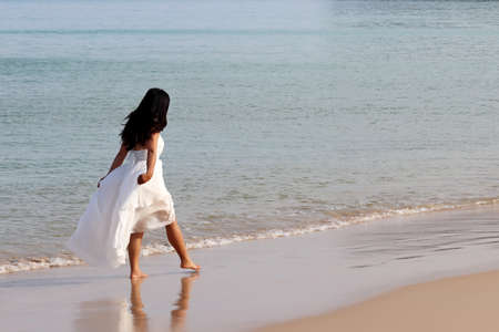 Woman in a white dress walking on a beach on background of morning sea, happy traveler. Concept of honeymoon, vacation and travel, romance and freedom, wedding fashionの写真素材