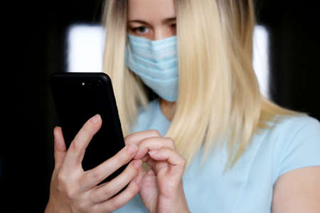 Blonde woman in face mask using smartphone, mobile phone in female hands closeup. Concept of safety...