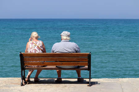 Elderly woman and man sitting on a wooden bench on blue sea background, rear view. Old couple on a beach, relax and enjoying life in retirementの写真素材