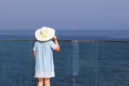 Woman in sun hat and blue dress standing on the glass terrace on sea background. Beach relax and vacation, enjoying life conceptの写真素材
