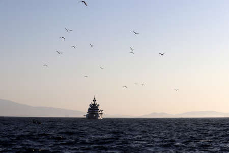 Picturesque seascape with luxury yacht and flying seagulls silhouettes on misty mountains background. Scenic view to the evening sea and islandsの写真素材