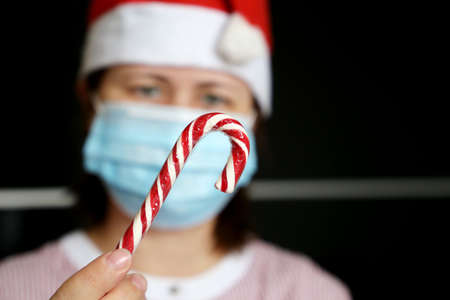 Portrait of woman in medical face mask and Santa Claus hat with lollipop in hand. Christmas celebration during coronavirus pandemicの写真素材