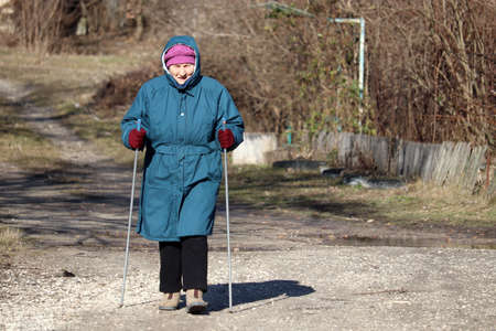 Elderly woman walking with sticks on rural road, sports exercises for healthy spine and joints. Nordic walking at cold weather, therapy for healthの写真素材