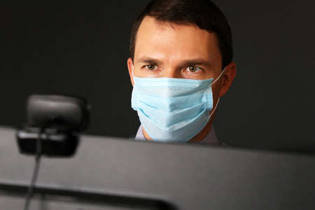 Man in face mask during online video chat in front of a webcam and PC monitor. Office worker, safety...