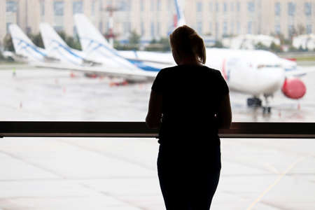 Silhouette of woman in the airport terminal, selective focus. Passenger waiting for their flight and looking to the airplanes through the window glassの写真素材