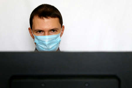 Man in face mask sitting at the PC monitor. Safety at office during coronavirus pandemic