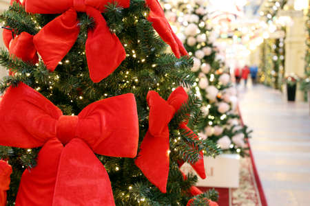 Christmas tree with red festive bows in a shopping mall on festive lights and walking people background. New Year decorations, sale on winter holidaysの写真素材