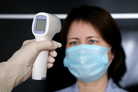 Coronavirus symptoms, doctor measures body temperature of woman in medical mask. Digital infrared...