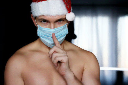 Portrait of muscular man in medical face mask and Santa Claus hat asking to be quiet with finger on lips. Christmas celebration during coronavirus pandemicの写真素材