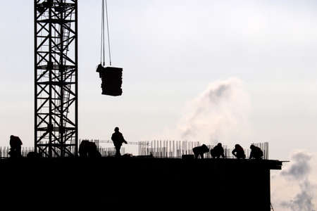 Silhouettes of workers and construction crane with cargo against the sky. Housing construction,...