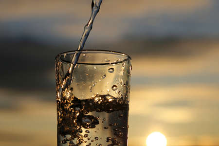 Pouring clean water into drinking glass on sunset blurred background, selective focus. Concept of health and freshness, thirst, water purificationの写真素材