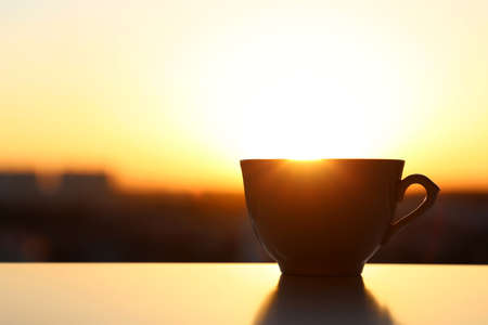 Silhouette of coffee cup on sunrise background, fresh start in the morning. Cozy atmosphere, city view from the windowの写真素材