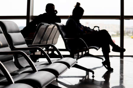 Passengers in the airport, defocused silhouettes of man and woman sitting against the window. Travelers waiting flightの写真素材