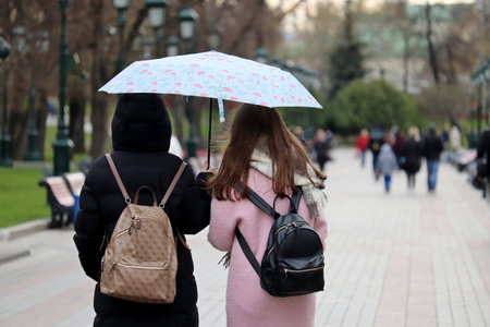 Rain in city, two girls with one umbrella walk on a street on people background. Rainy weather, spring stormのeditorial素材
