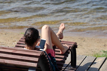 Girl in shorts sitting on deck chair with smartphone on a beach, mobile phone in female hands. Concept of using phone in travel, online communication at the seasideの写真素材