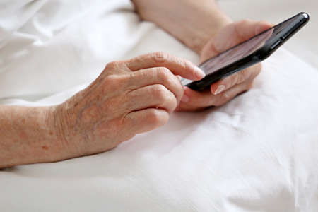 Elderly woman with smartphone sitting in bed, mobile phone in wrinkled female hands close up....