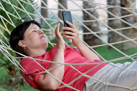 Woman relaxes in a hammock with smartphone in hands on palm trees background. Summer leisure in the...