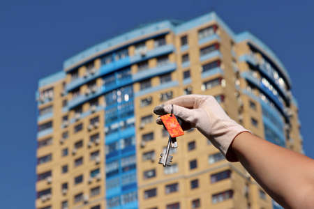 House purchase, woman in protective gloves holding keychain with digital and home keys on background of new buildings. Moving home or renting property during coronavirus pandemicの写真素材