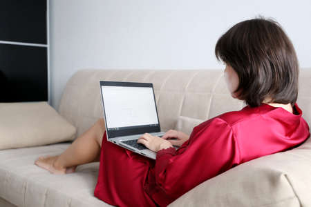 Woman with laptop sits on sofa, remote work during quarantine at home. Girl in red nightie typing on...