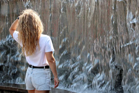 Blonde girl in jeans shorts with curly long hair standing against the water jets of fountain. Summer...