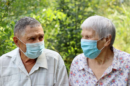 Elderly couple in protective face masks looking at each other on nature background. Safety during coronavirus pandemic, life in retirementの写真素材