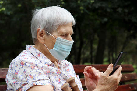 Elderly woman in protective mask sitting with smartphone on a bench in park. Safety during...