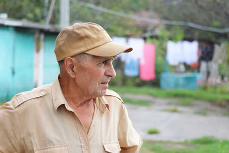 Elderly man standing in rural yard on background of well and hanged laundry. Concept of life in village, old ageの写真素材