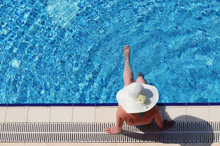 Woman in bikini and sun hat sitting on the edge of swimming pool and dangling her legs in water, top view. Vacation on beach resort, tanning and leisure conceptの写真素材