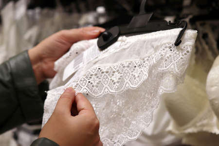 Woman chooses white lace panties in lingerie store, selective focus on hands. Female fashion, different sizes of wedding underwearの写真素材