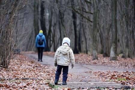 Little boy stands in autumn park on background of departing woman. Concept of offended child, orphan, parents divorceの写真素材