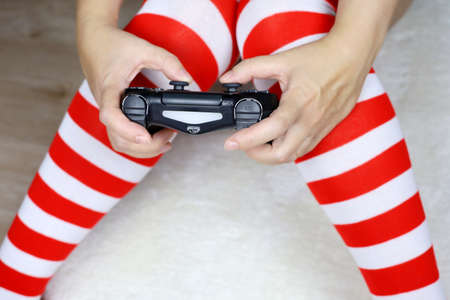 Woman gamer in Christmas knee socks playing video games with gamepad sitting on white fur rug. Home...