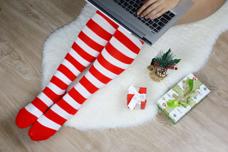 Woman in Christmas knee socks sitting on a fur rug with laptop near the gift boxes. Ordering gifts, work and leisure at home during New Year holidaysの写真素材