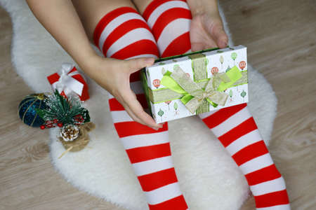 Girl in Christmas knee socks opens gift box sitting on white fur rug. Romantic present for New Year...