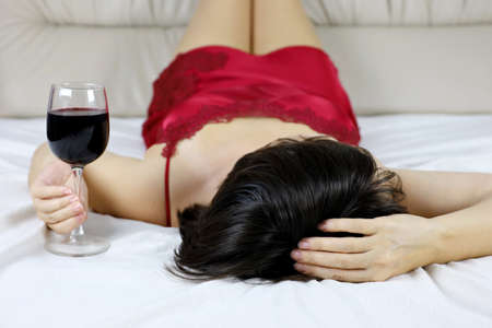 Woman in lingerie lying with glass of red wine on a bed. Concept of celebration, relaxation, female alcoholismの写真素材