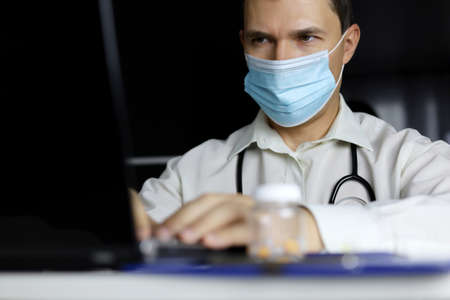 Doctor in disposable mask working at laptop in office. Physician writes prescription, concept of...