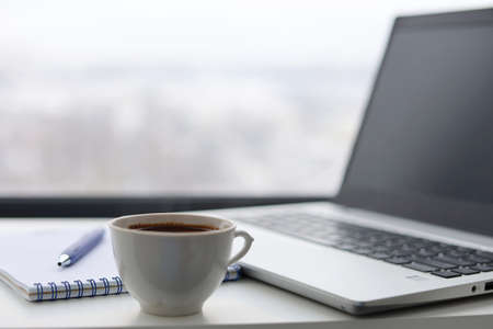 Coffee cup, laptop and notepad with pen on a table against the window, view to city. Cozy workplace...