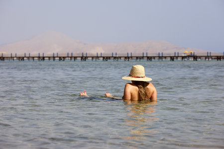Woman in sun hat sitting in sea water. Sun tan and vacation on summer beachの写真素材