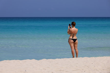 Woman in swimsuit taking pictures of blue sea on smartphone camera standing on a tropical sandy...
